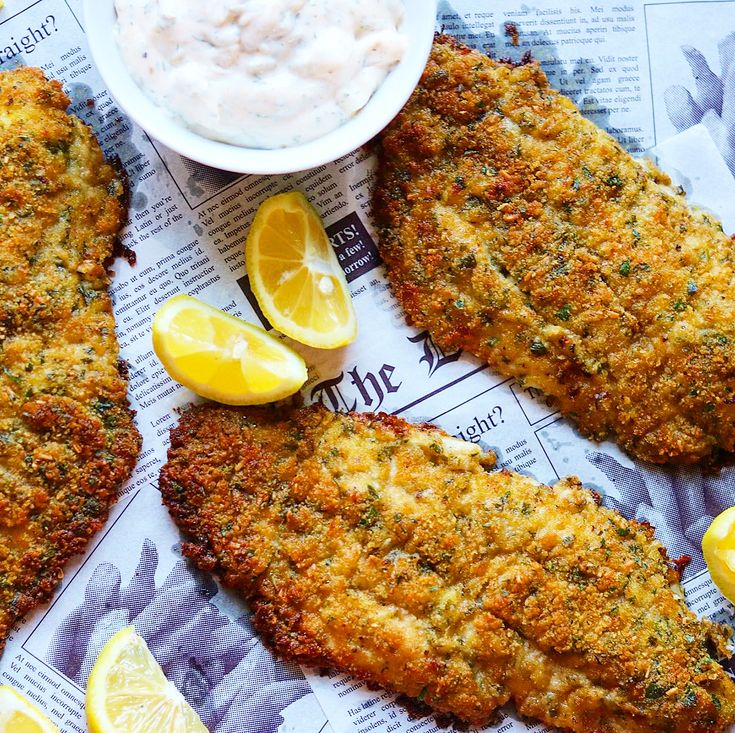 two fish fillets with lemon wedges on top of newspaper next to a bowl of dip