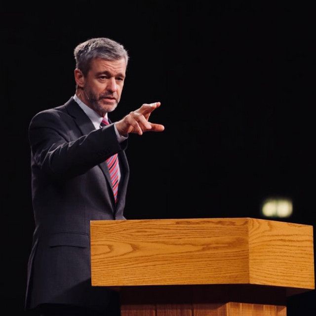 a man in a suit and tie giving a speech at a podium with his hand out