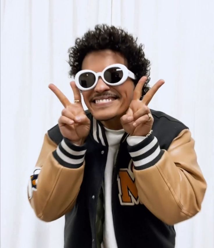 a man wearing sunglasses and a jacket making the peace sign with his hands while standing in front of a white curtain