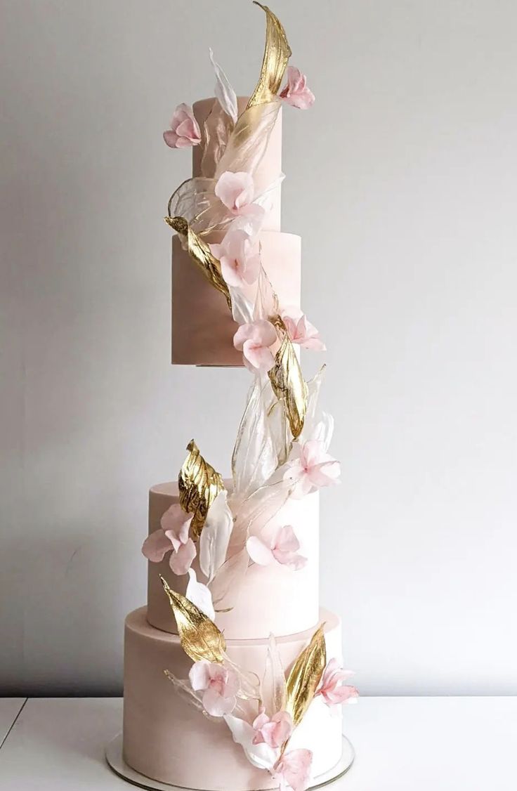 a three tiered cake with pink flowers and gold leaves on the top, against a white background