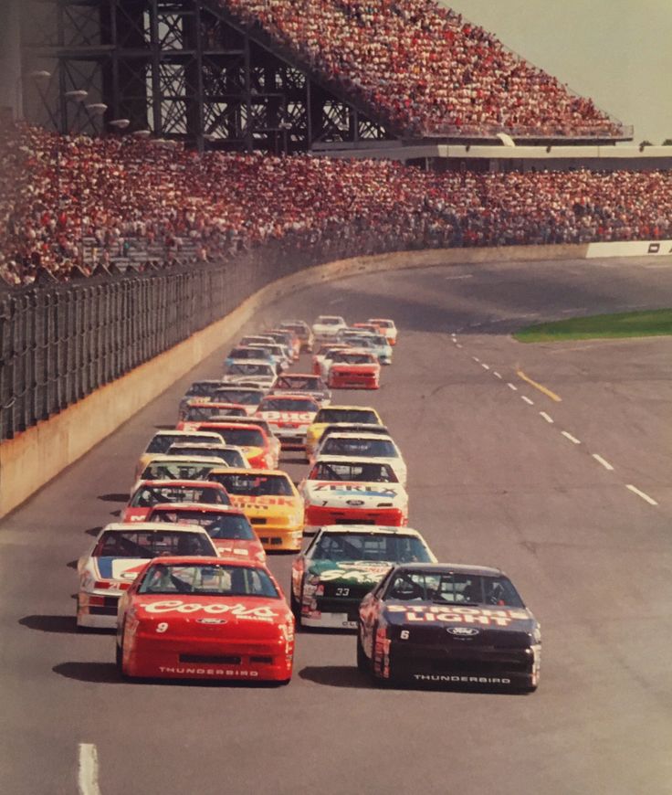 a group of cars driving down a race track in front of a crowd of people