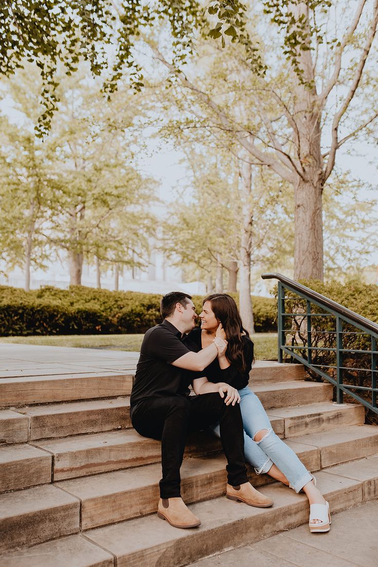 an engaged couple sitting on the steps in front of some trees and stairs with their arms around each other