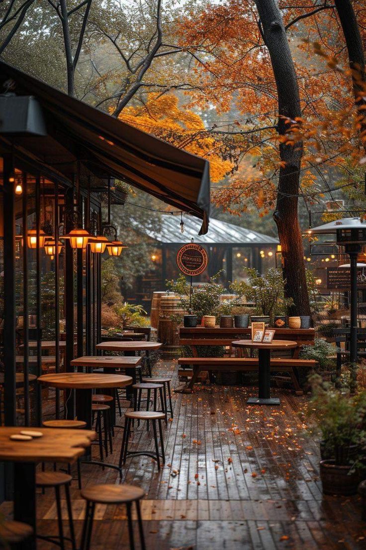 an outdoor restaurant with tables and umbrellas in the rain, surrounded by trees that have orange leaves on them