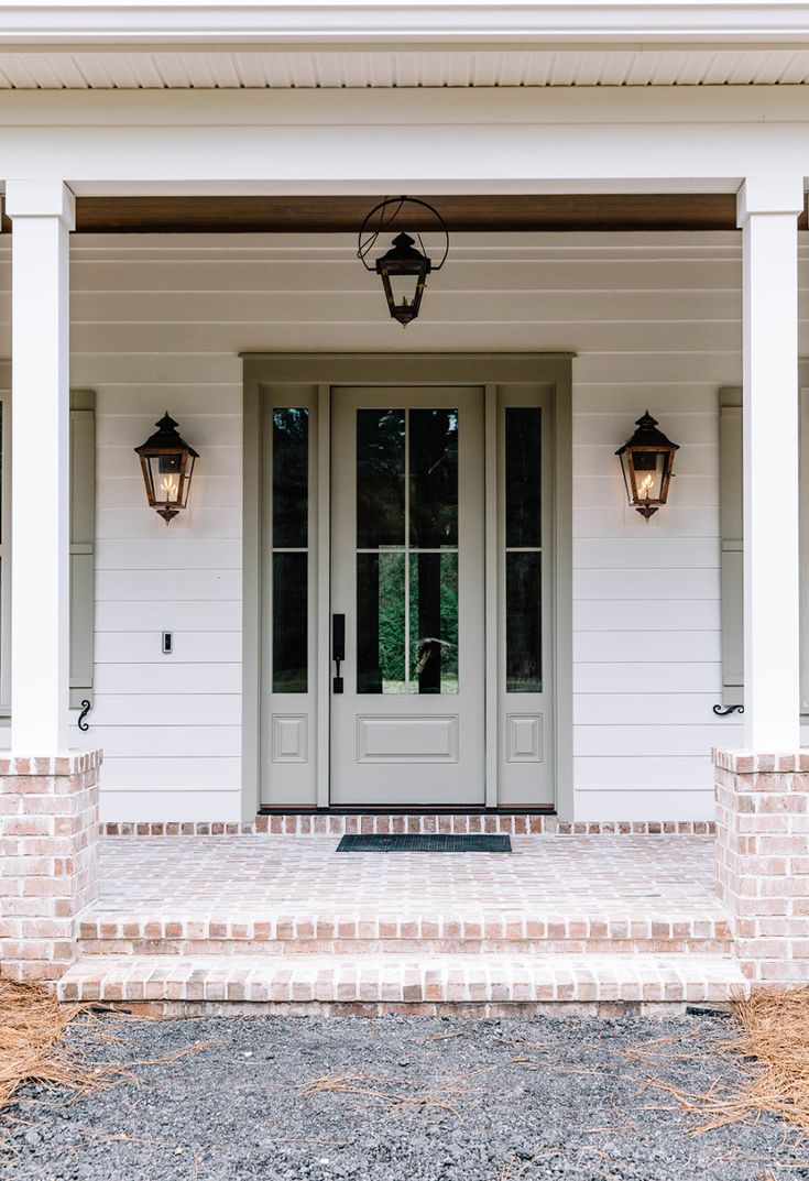 the front entrance to a white house with two lights on each side and brick pillars