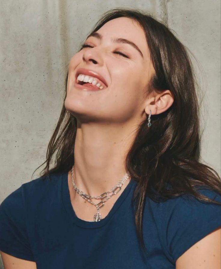 a woman smiling with her eyes closed in front of a concrete wall and wearing a blue t - shirt