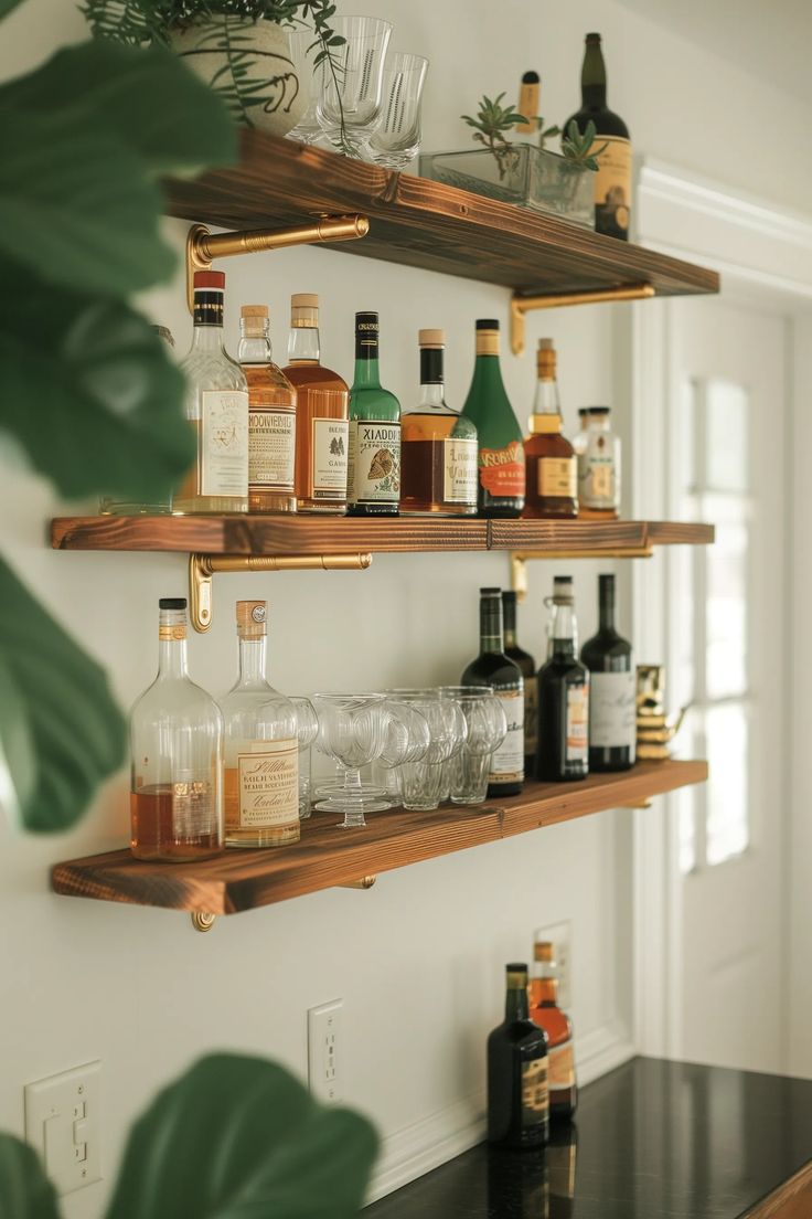 several bottles and glasses on wooden shelves in a room with white walls, black countertops and green plants