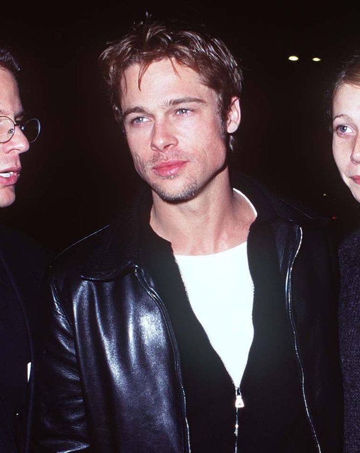 three people standing next to each other in front of a building and one man is wearing a black leather jacket