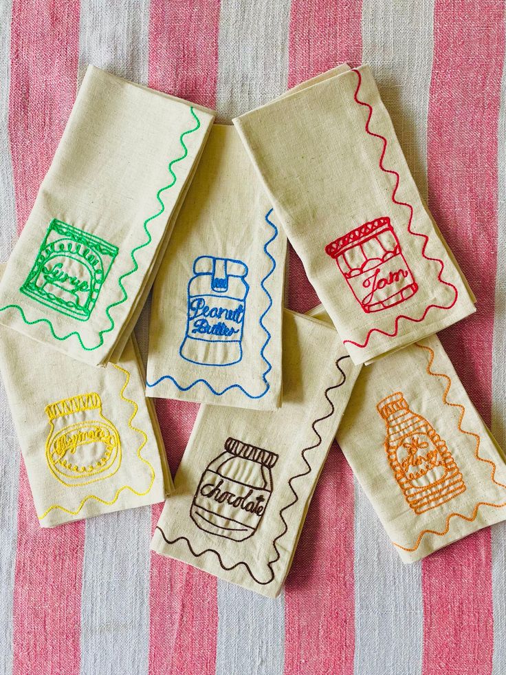 six hand embroidered tea towels on a pink and white striped tablecloth with different designs