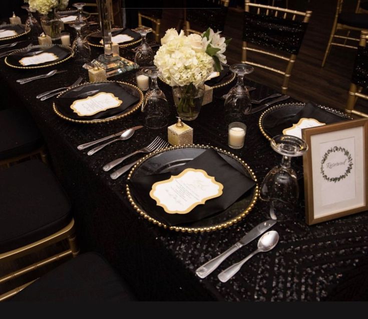 a table set with black and gold place settings, silverware, and white flowers