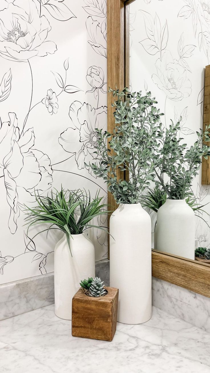 two white vases with plants in front of a mirror on a marble countertop