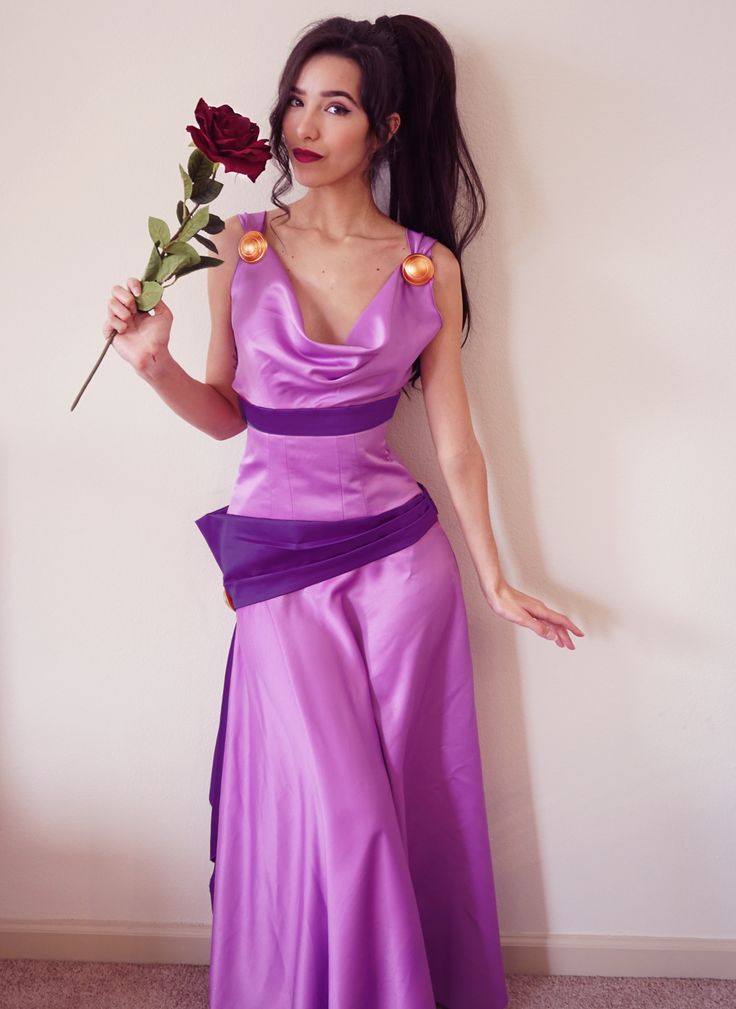 a woman in a long purple dress holding a rose and looking at the camera while standing against a wall