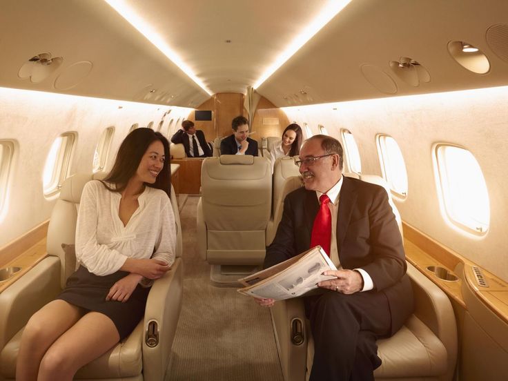 two people are sitting on an airplane and one person is reading a newspaper while the other man looks at him