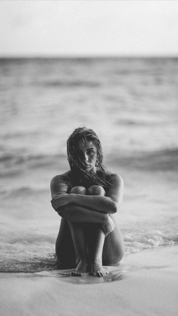 a woman is sitting on the beach with her arms around her chest and looking at the water