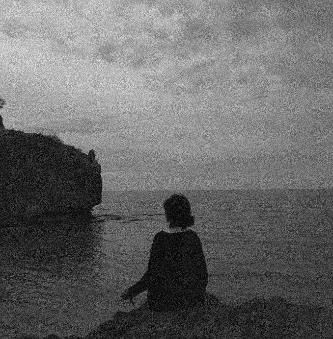 black and white photograph of person sitting on rocks near water