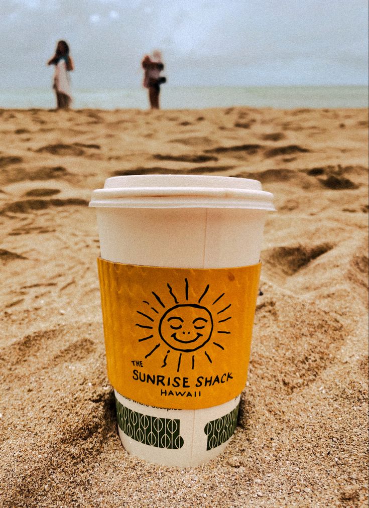 a cup of coffee sitting on top of a sandy beach next to the ocean with two people in the background