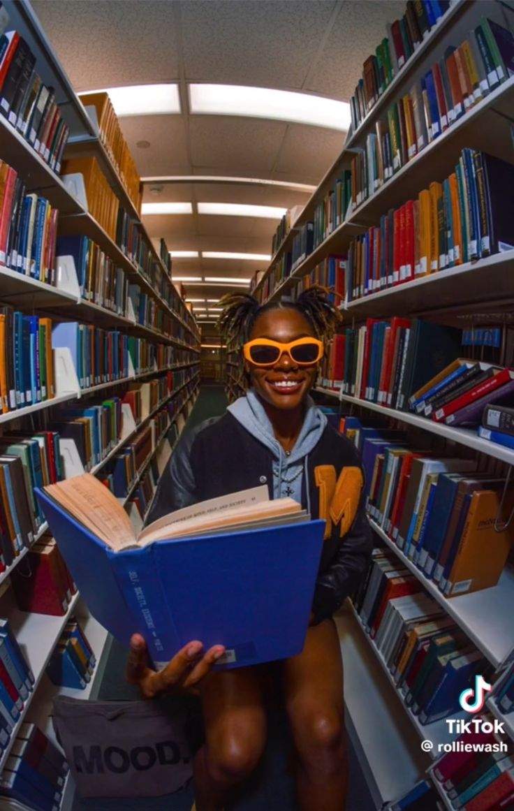 a woman sitting in the middle of a library holding a book and smiling at the camera