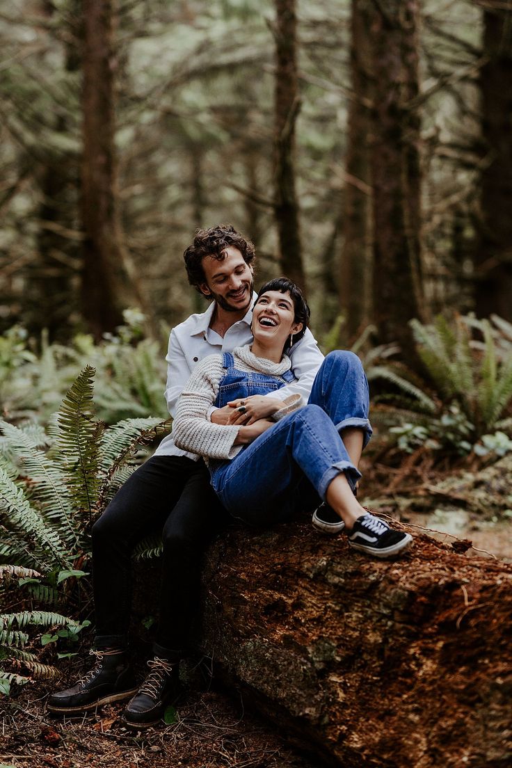 a man and woman are sitting on a log in the woods smiling at each other