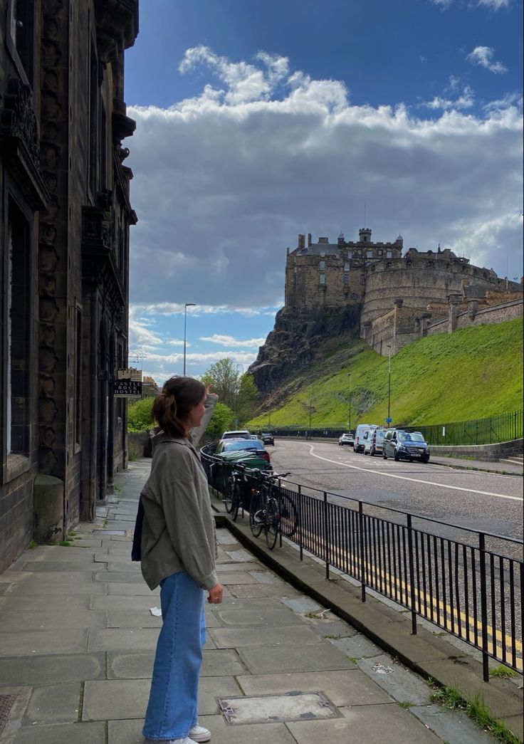 a woman is standing on the sidewalk looking at cars parked in front of a castle