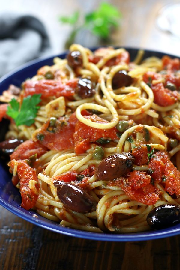 a blue bowl filled with pasta, olives and tomato sauce on top of a wooden table