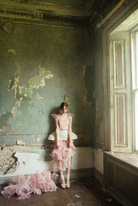 a woman in a pink dress standing next to a bathtub with peeling paint on the walls