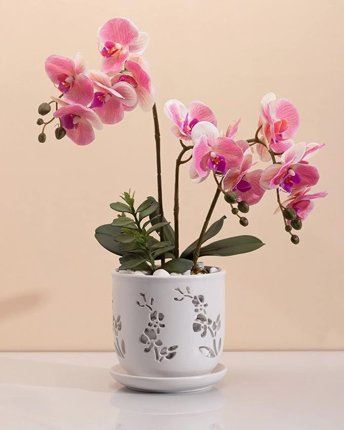 pink orchids are in a white vase on a table with a beige wall behind them