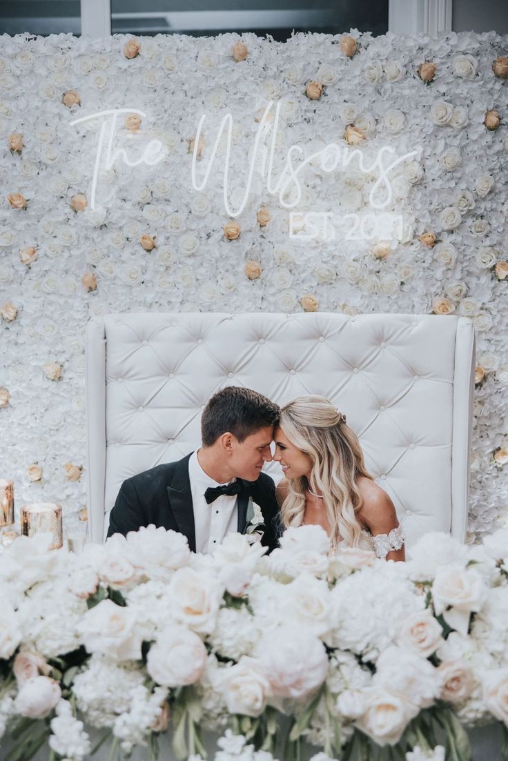 a bride and groom sitting in front of a floral backdrop at their wedding reception table