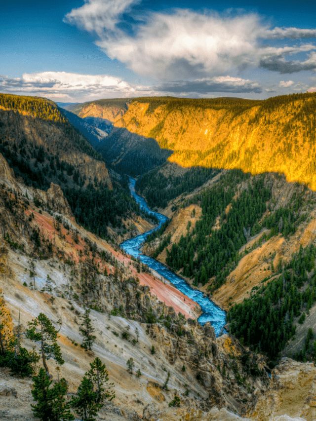 a river flowing through a canyon surrounded by trees and mountains in the distance with clouds overhead