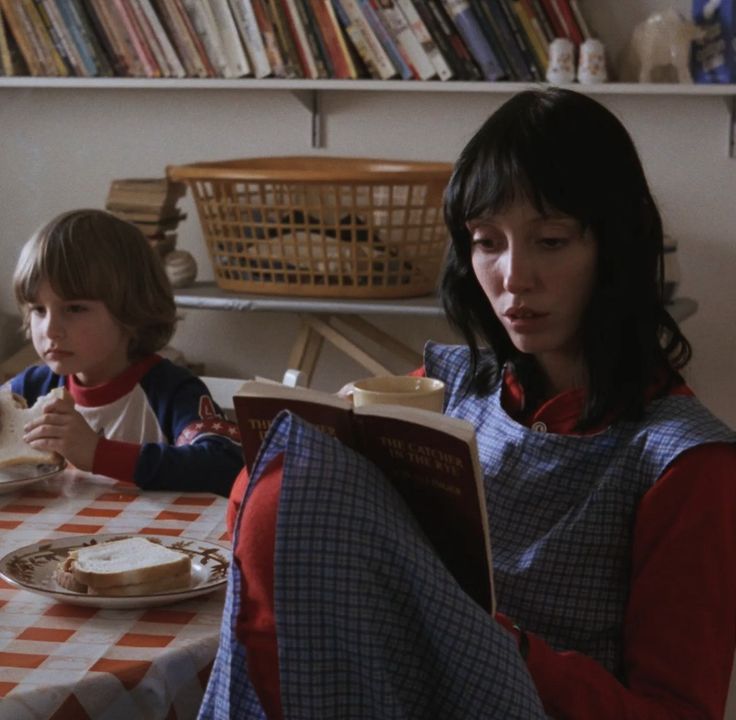 two children sitting at a table with food in front of them and one reading a book