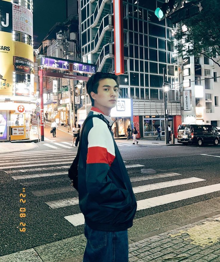 a young man standing on the side of a street at night with buildings in the background