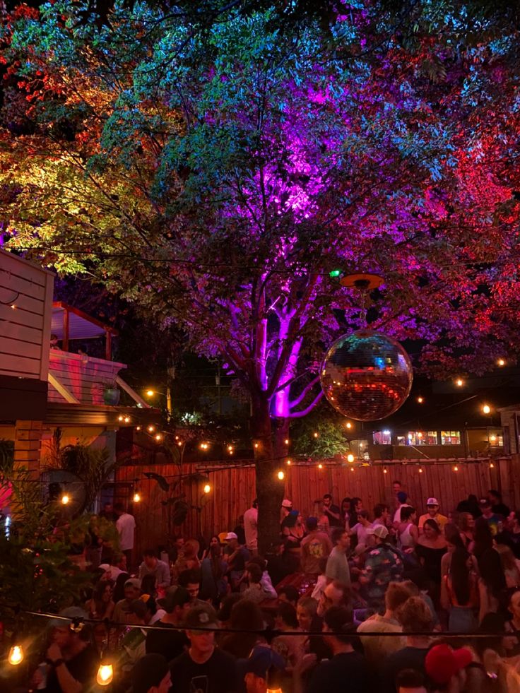 a group of people sitting around in front of trees with colorful lights on them at night