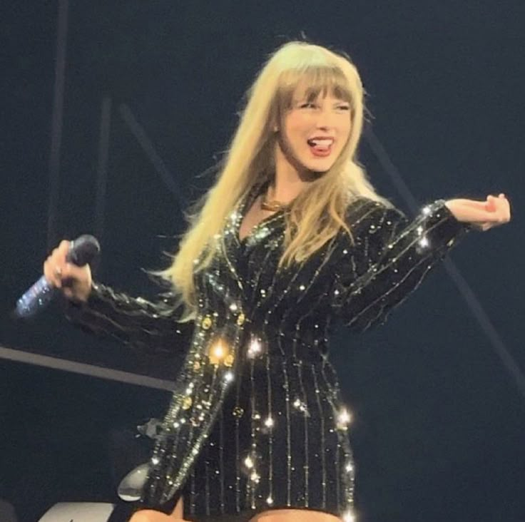 taylor swift performing on stage with microphone in her hand and sparkling outfit, holding out her right arm