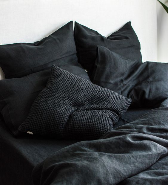 a bed with black linens and pillows on top of it, next to a plant