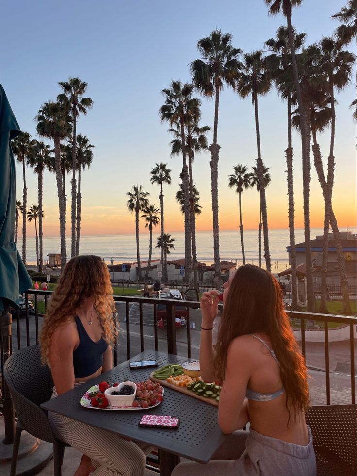 two women sitting at an outdoor table with food and drinks in front of palm trees