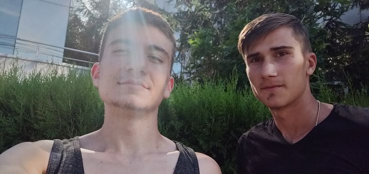 two young men standing next to each other in front of some bushes and trees with the sun shining on them