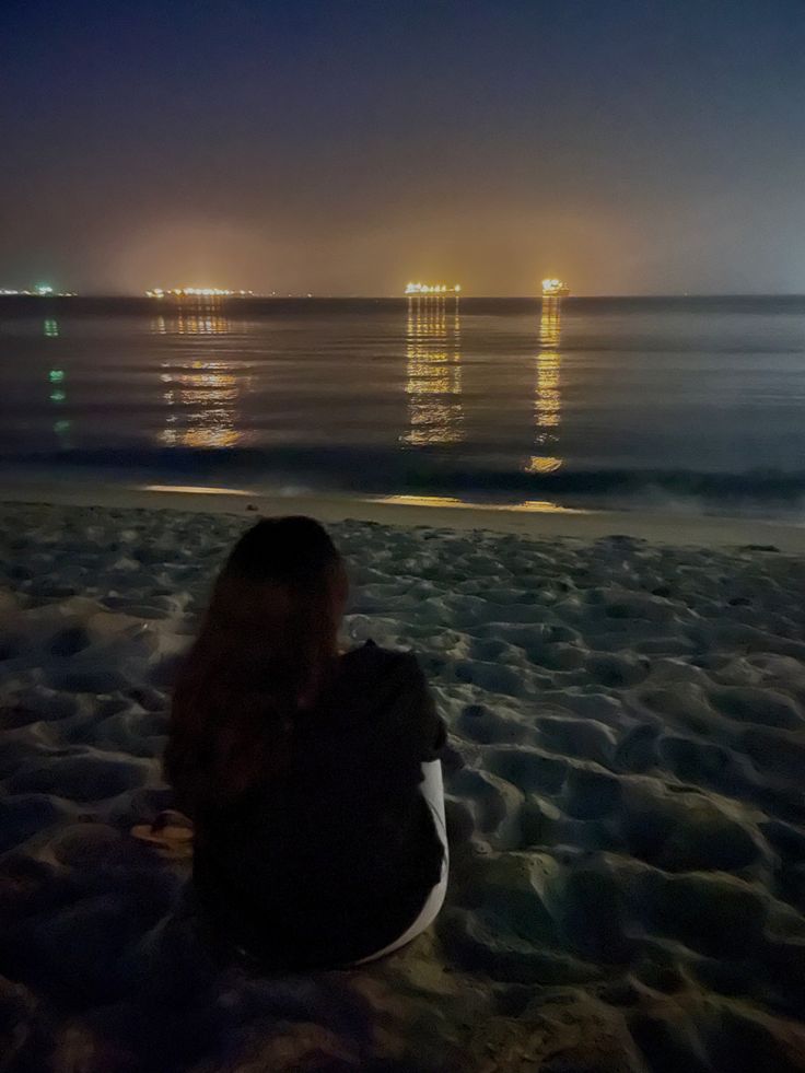 a woman sitting on the beach at night looking out to sea with lights in the distance