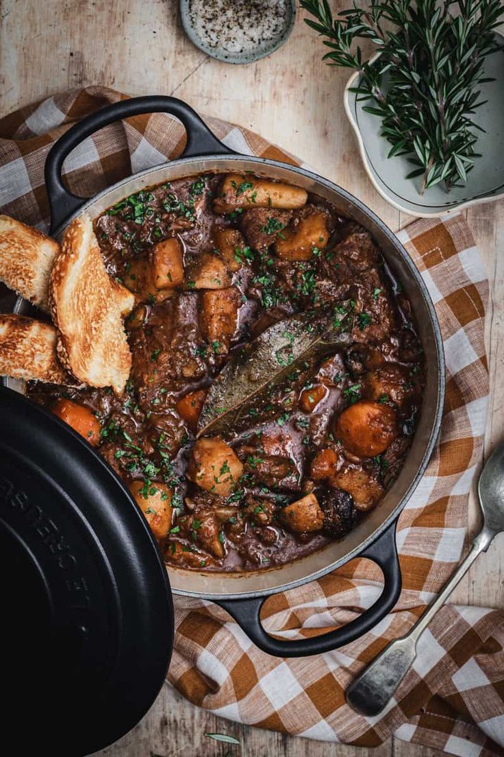 a pot filled with stew and bread on top of a wooden table next to utensils