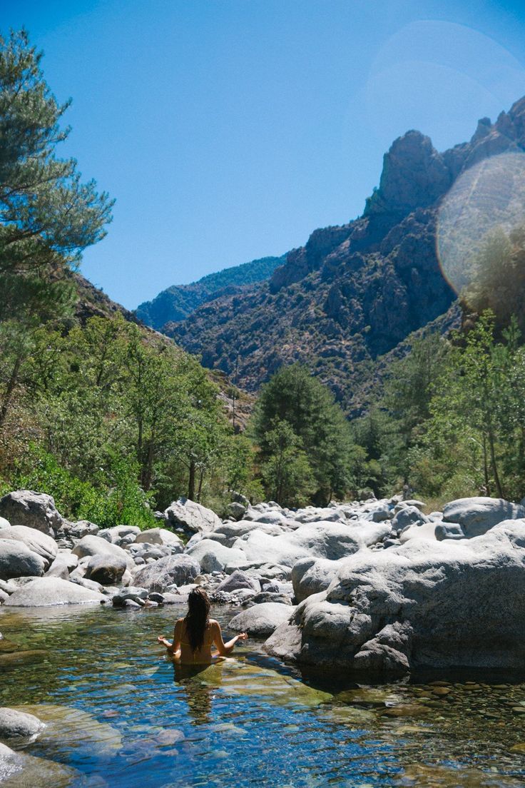 a woman is sitting in the middle of a river with rocks and trees around her