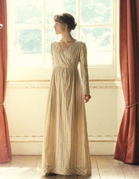 a woman standing in front of a window wearing a long beige dress with pleated sleeves