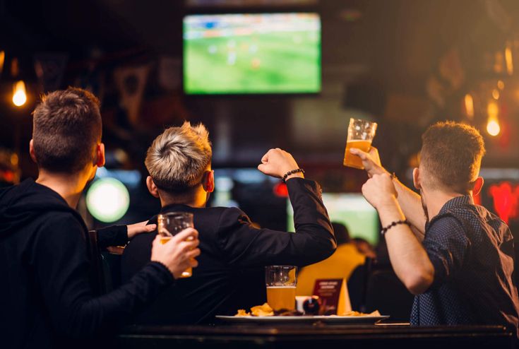 three men are sitting at a bar drinking beer and watching televisions in the background