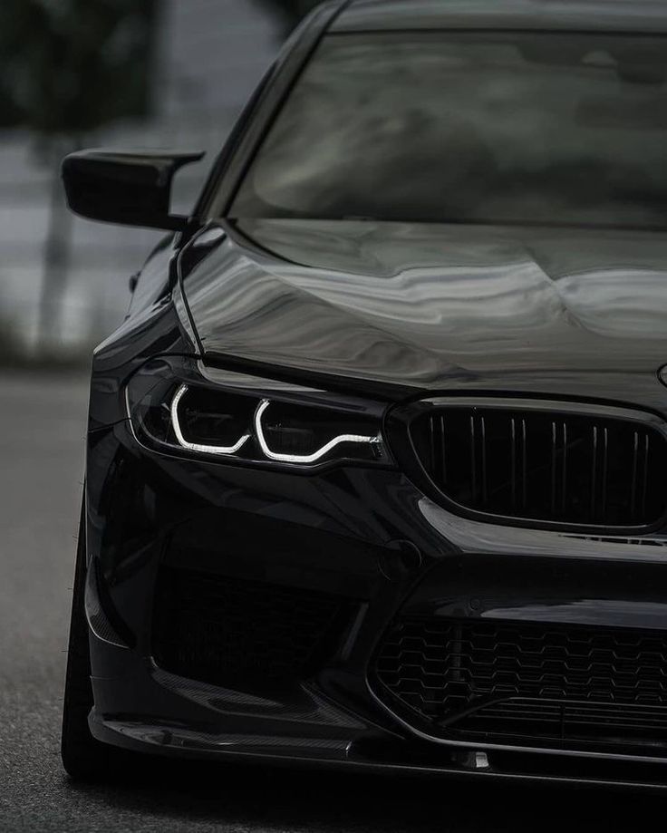 the front end of a black car parked in a parking lot with its lights on