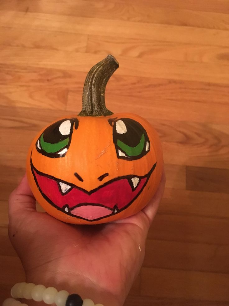 a hand holding an orange painted pumpkin with green eyes and fangs on it's face