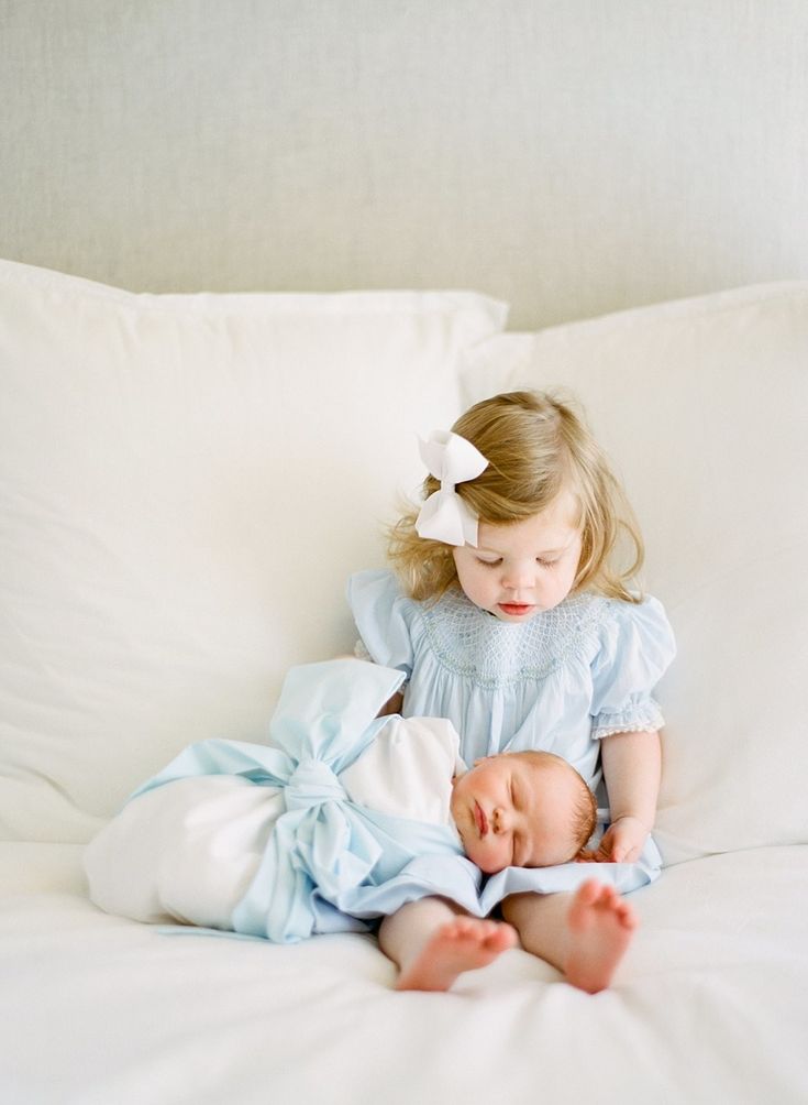 Image of a toddler wearing a blue dress, sitting on a bed with her newborn baby brother laying on her lap. He's wrapped in a blue bow swaddle. Blue And White Newborn Family Pictures, 2 Under 2 Newborn Pictures, Newborn Baby With Siblings, New Sibling Photoshoot, Newborn Boy Photography With Siblings, Newborn Photos With Sister, Newborn With Toddler Photography, Newborn With Sister Photography, Newborn Pictures With Toddler