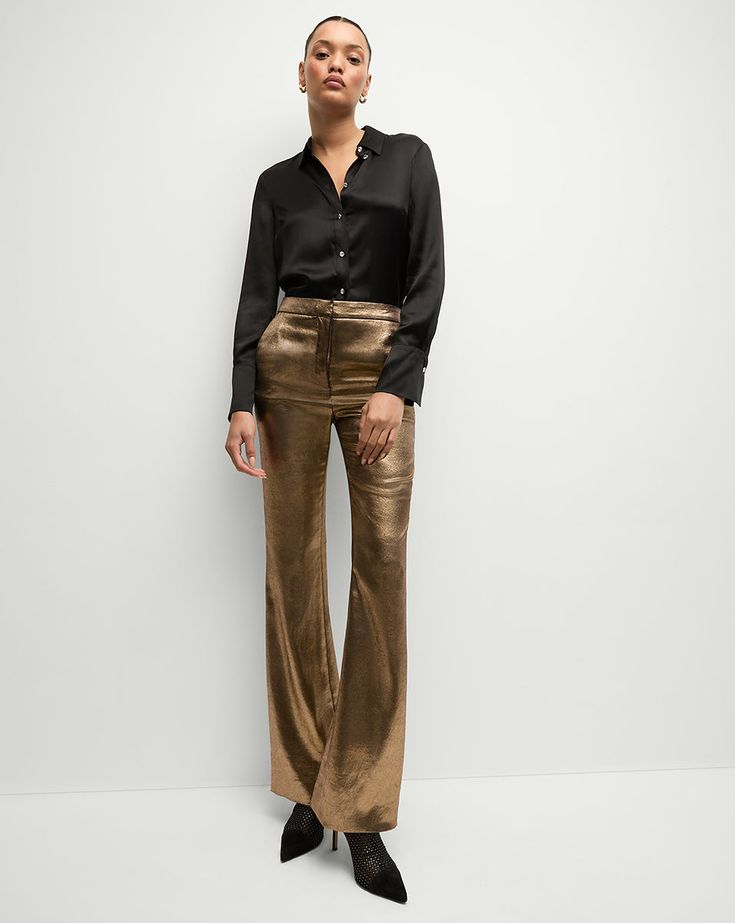 a woman standing in front of a white wall wearing gold pants and a black shirt