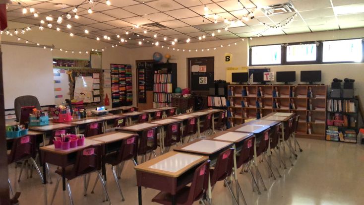 an empty classroom with desks and bookshelves in front of the windows, lights strung from the ceiling