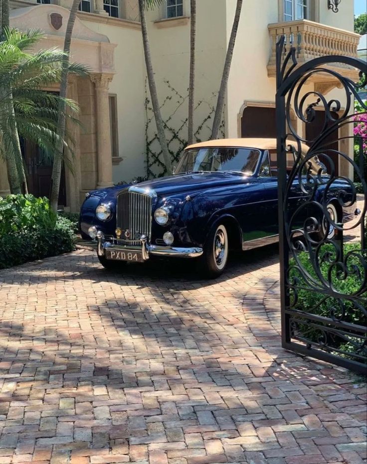 an old blue car parked in front of a house with palm trees and flowers on the driveway