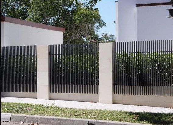 a black cat sitting on the side of a road next to a tall white fence