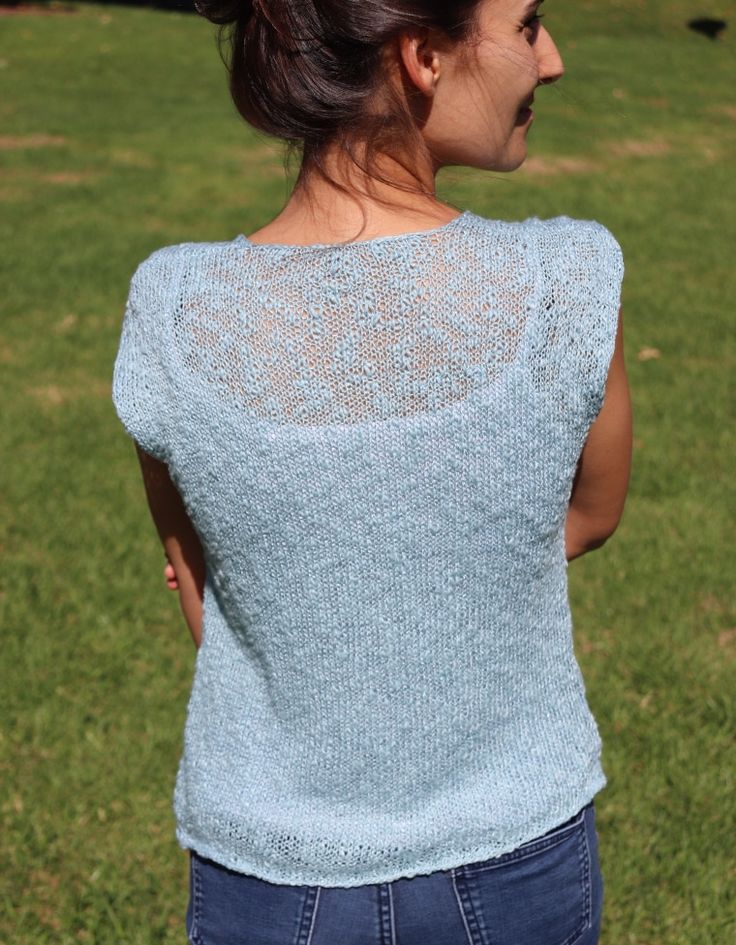 a woman standing in the grass with her back to the camera, wearing a light blue knitted top