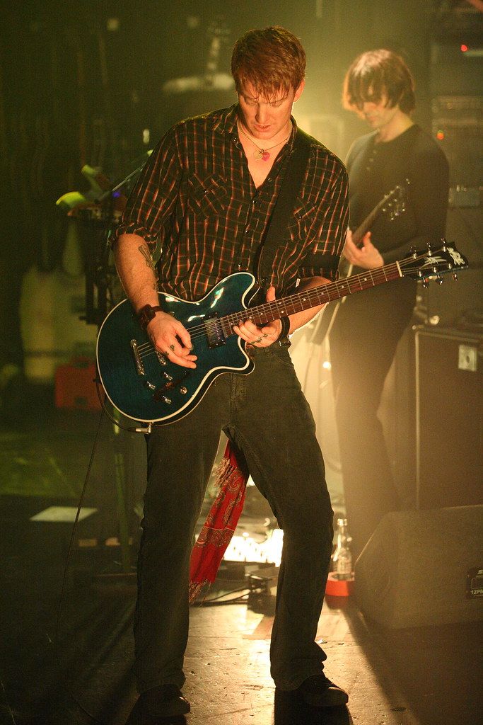 a man playing guitar on stage with another person in the background watching him play his instrument