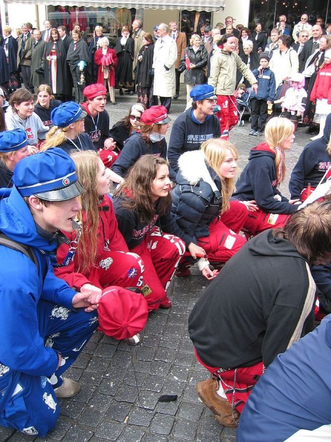 a group of people sitting on the ground in front of a building with many other people standing around