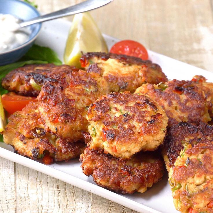 several crab cakes on a white plate with tomatoes and lemon wedges next to it